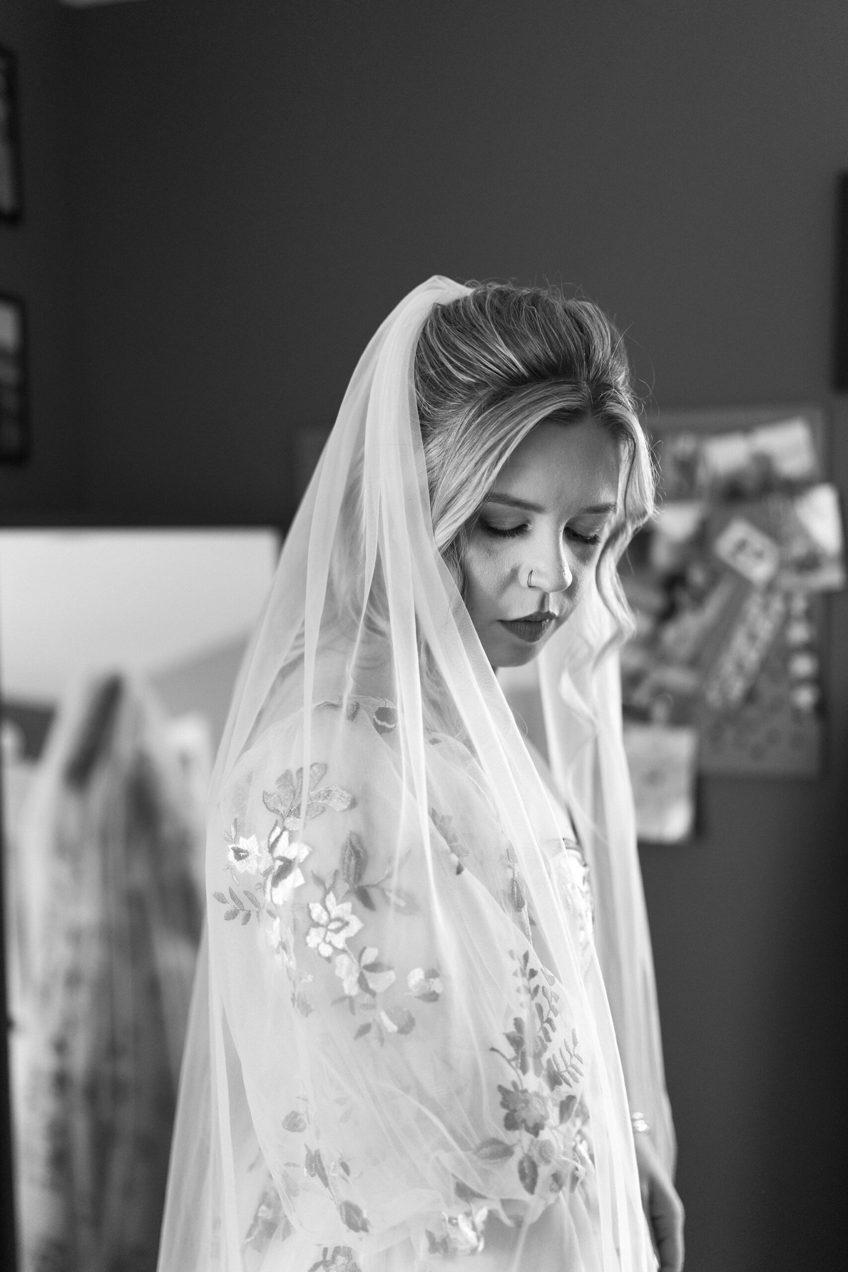 black and white image of a bride looking down over her right shoulder wearing an embroidered wedding dress, her veil wrapped around her shoulders.