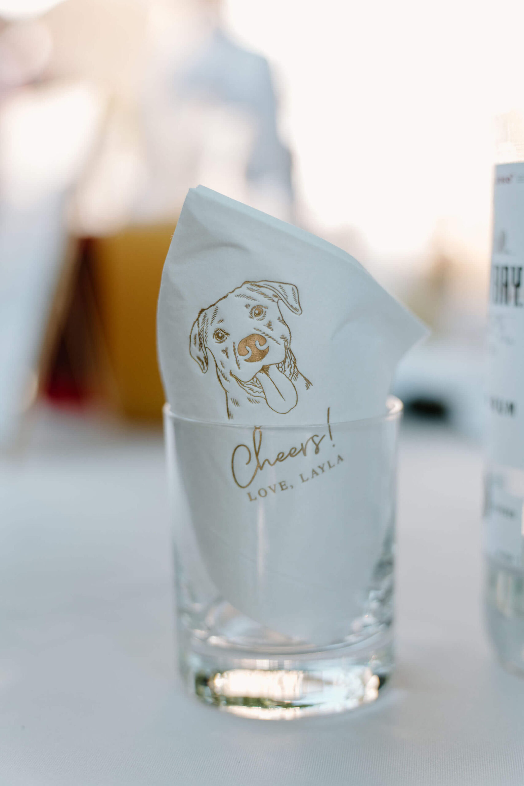 a napkin stamped with a picture of a happy-looking dog, inscribed with "Cheers! Love, Layla), propped inside a rocks glass