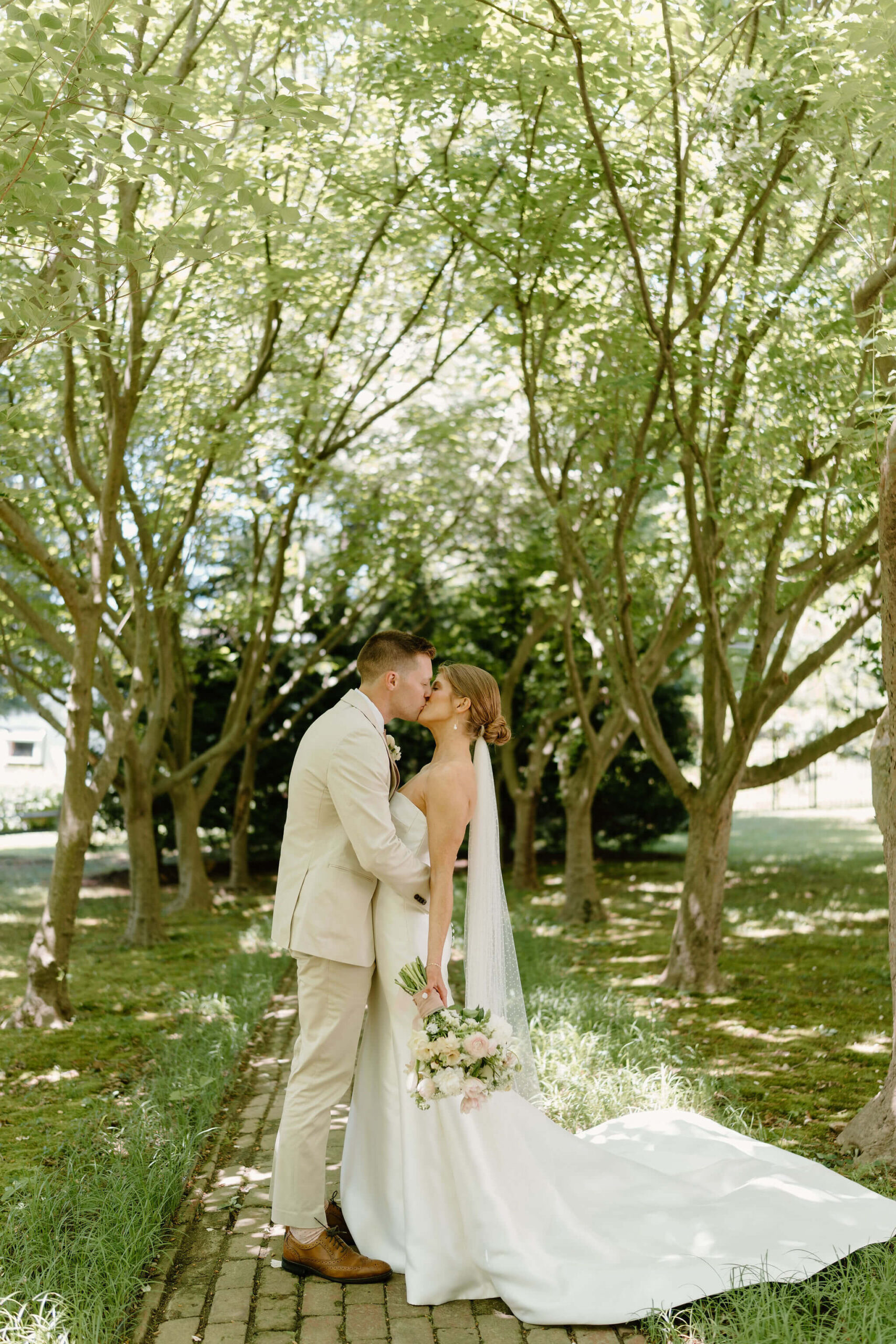 groom (tan suit, brown leather shoes) embracing and kissing his bride (low bun, white wedding gown, bridal bouquet down at her side) on a shaded path through trees