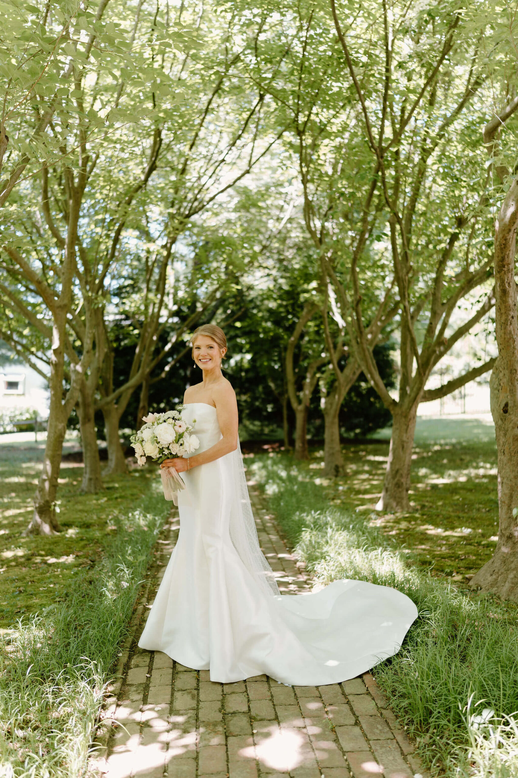 bride in a white wedding mermaid wedding gown smiling at the camera underneath a canopy of trees, holding a bridal bouquet