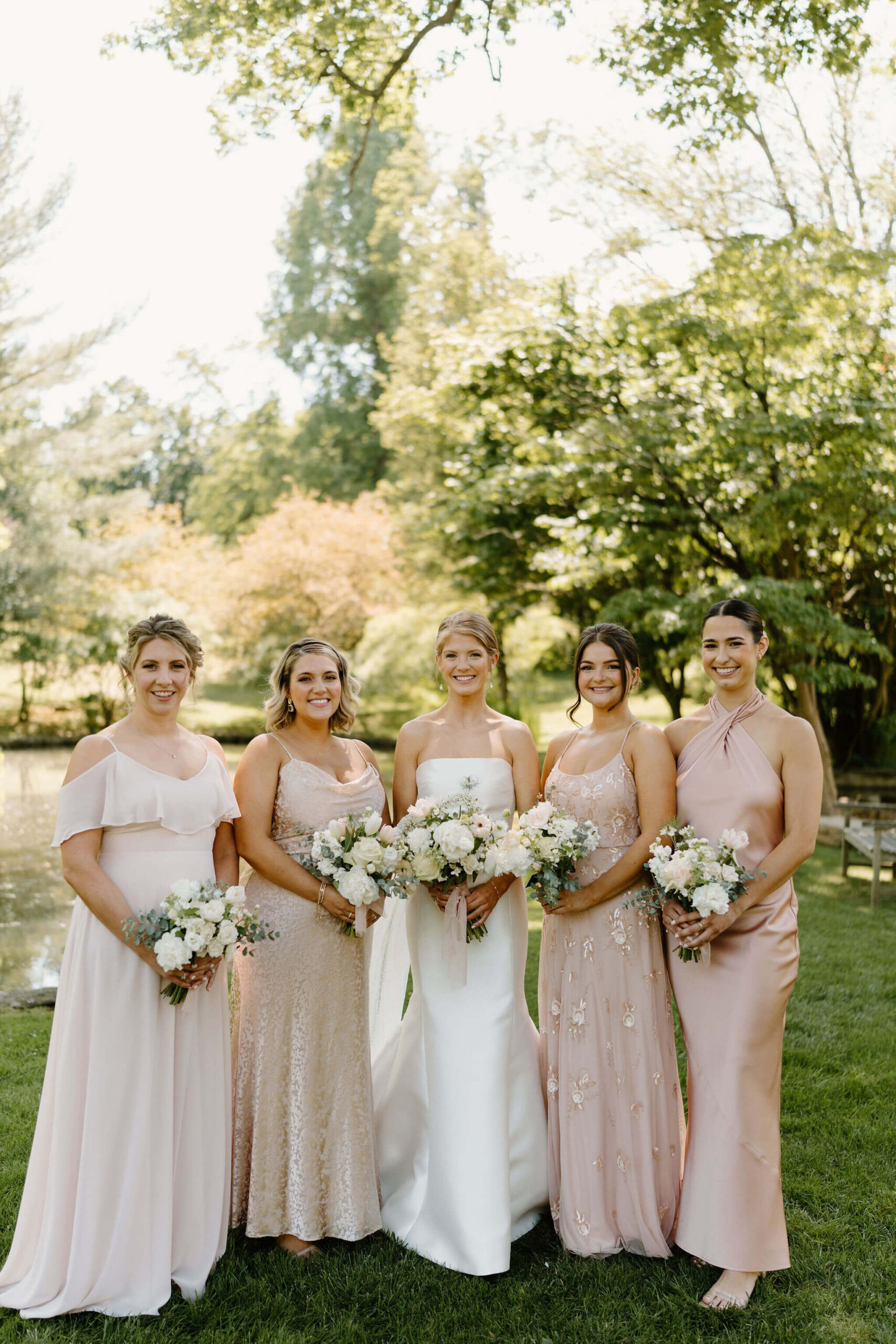 bride (white mermaid wedding gown) and bridesmaids (mismatched pink dresses) holding bouquets and smiling at the camera