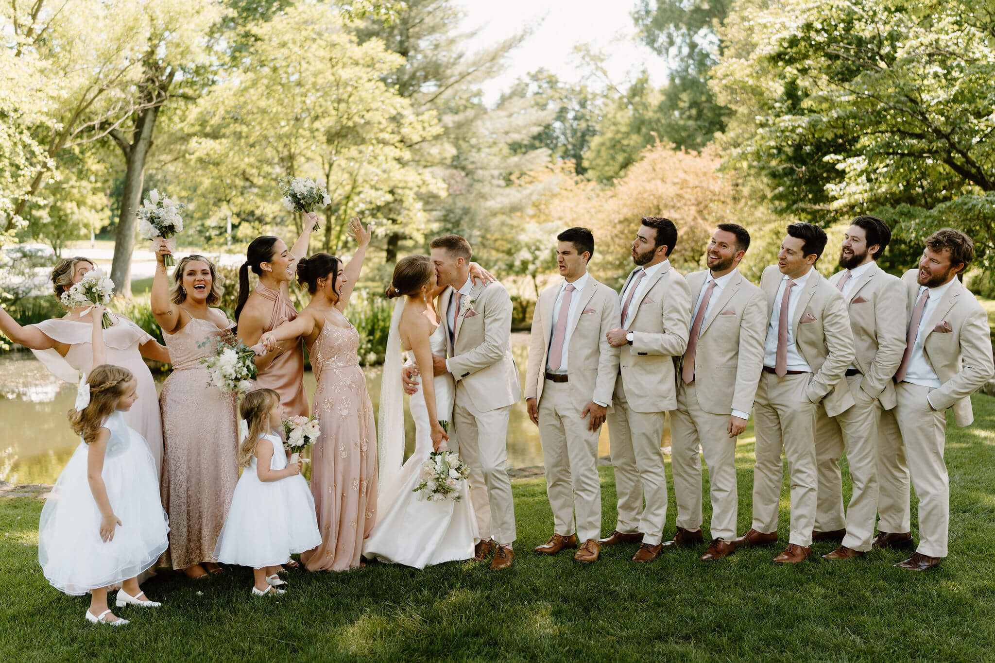 wedding party portrait of groomsmen in tan suits and pink ties, bridesmaids wearing mismatched dresses and throwing their hands in the air while holding bouquets, and two flower girls in white dresses, all smiling and looking toward the bride and groom kissing in the center of the group