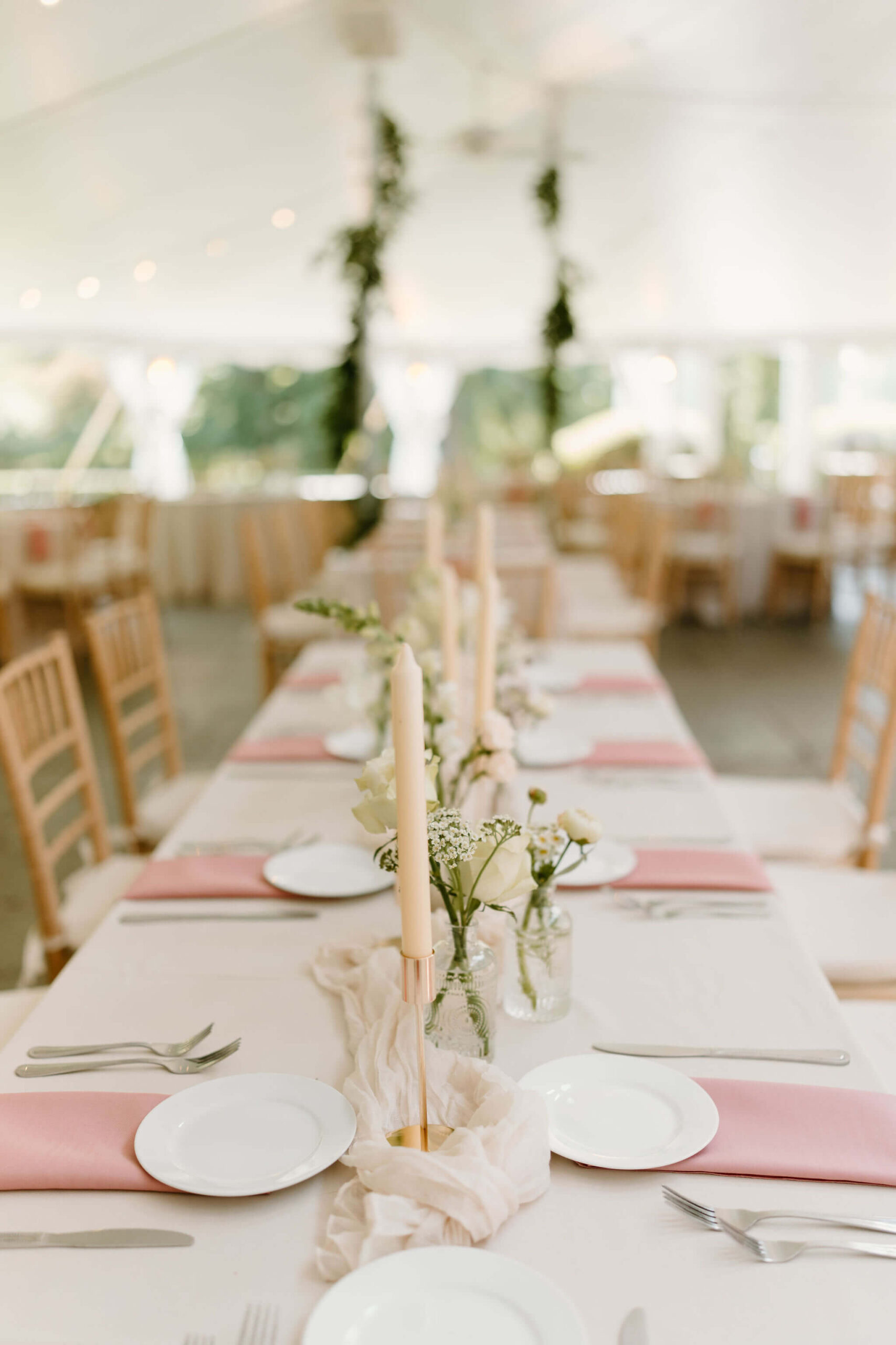 view down a long reception table with a white tablecloth, pink napkins, and tall candlesticks with small vases of white florals and greenery