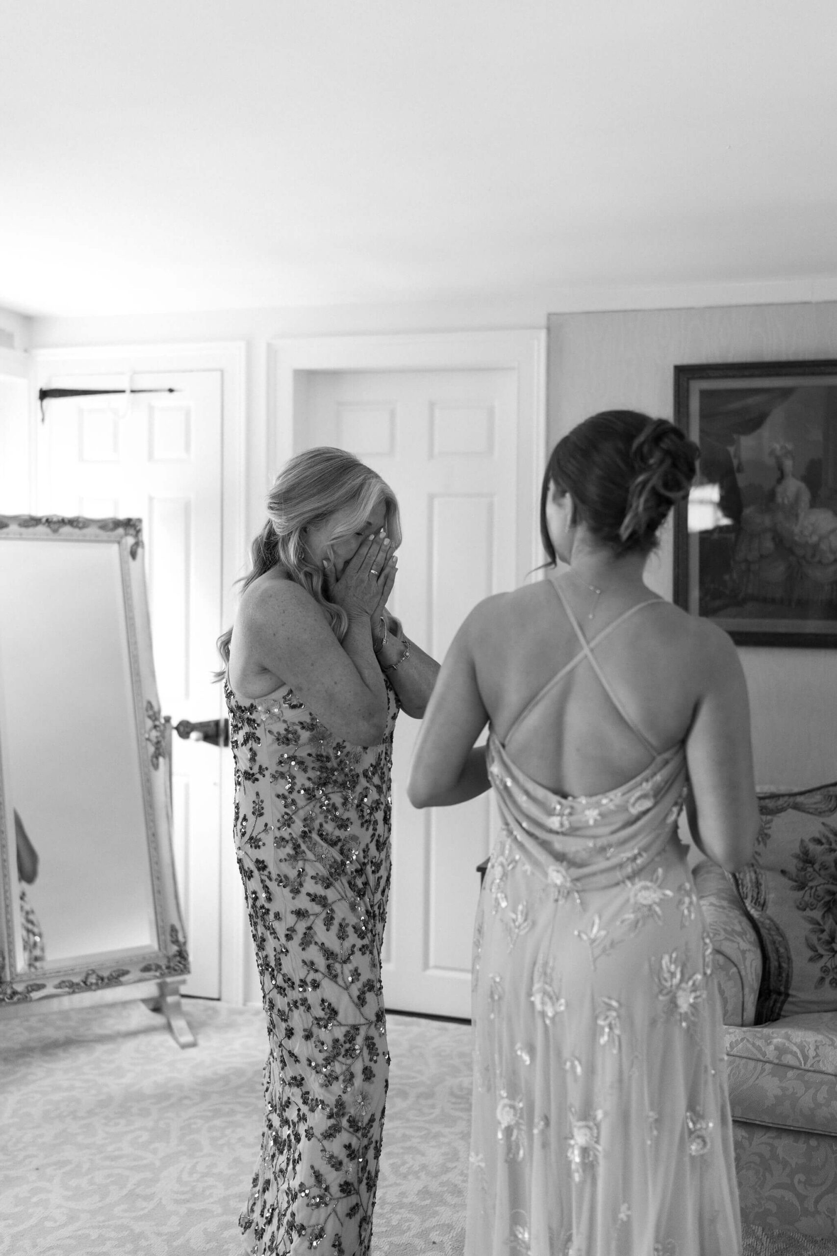 black and white image of the mother of the bride in a floral sequined dress, crying with her hands over her face while the sister of the bride looks toward her