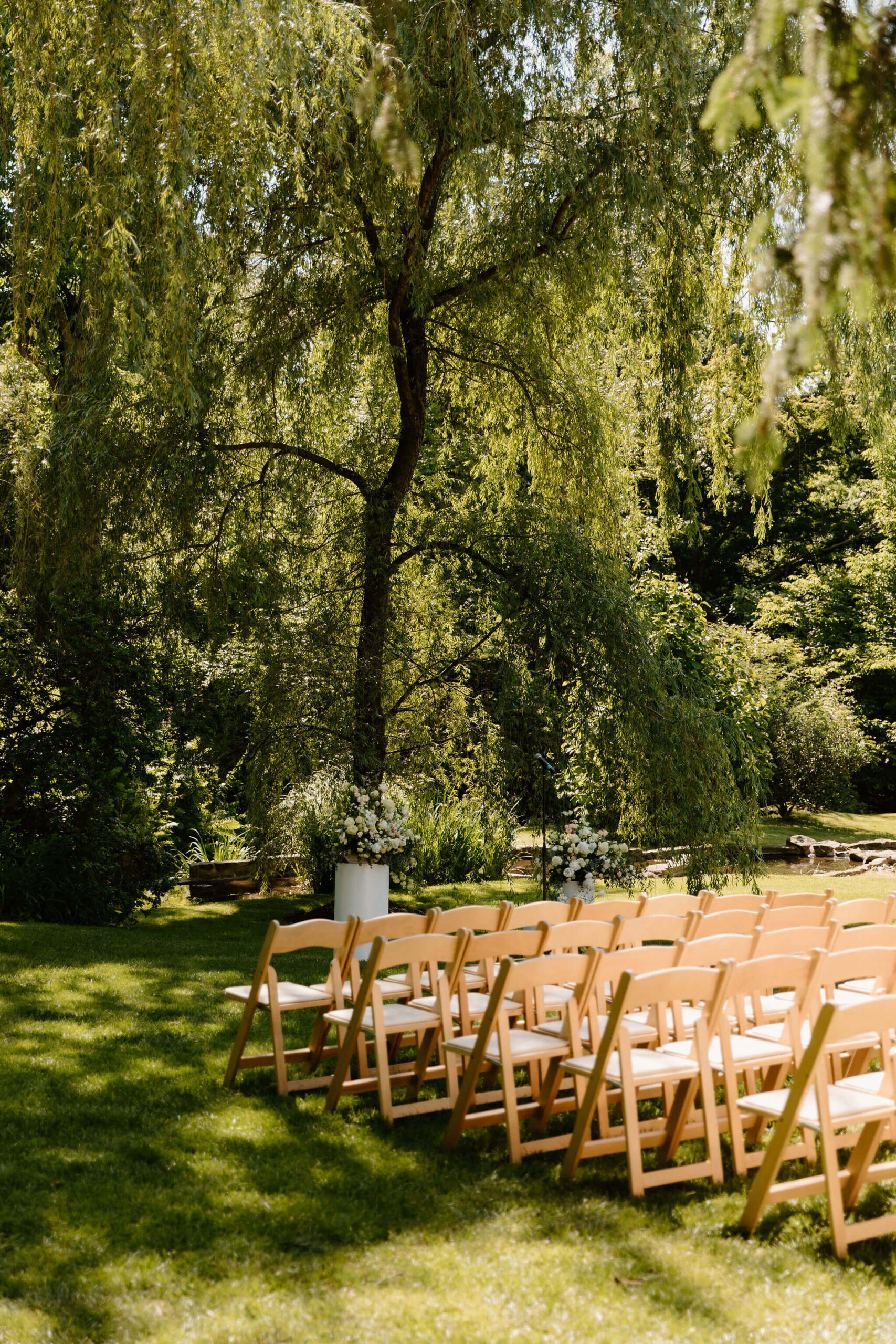light wooden ceremony chairs in a green and wooded area, with two floral centerpieces.