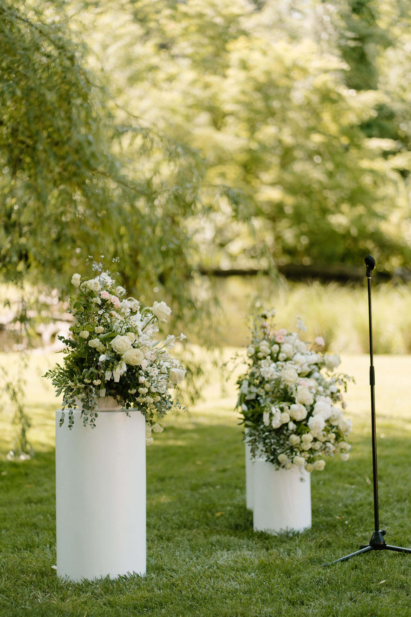 spring wedding ceremony decor of two floral arrangements with white flowers and lots of greenery, on two multi-level stands, against a lush green background