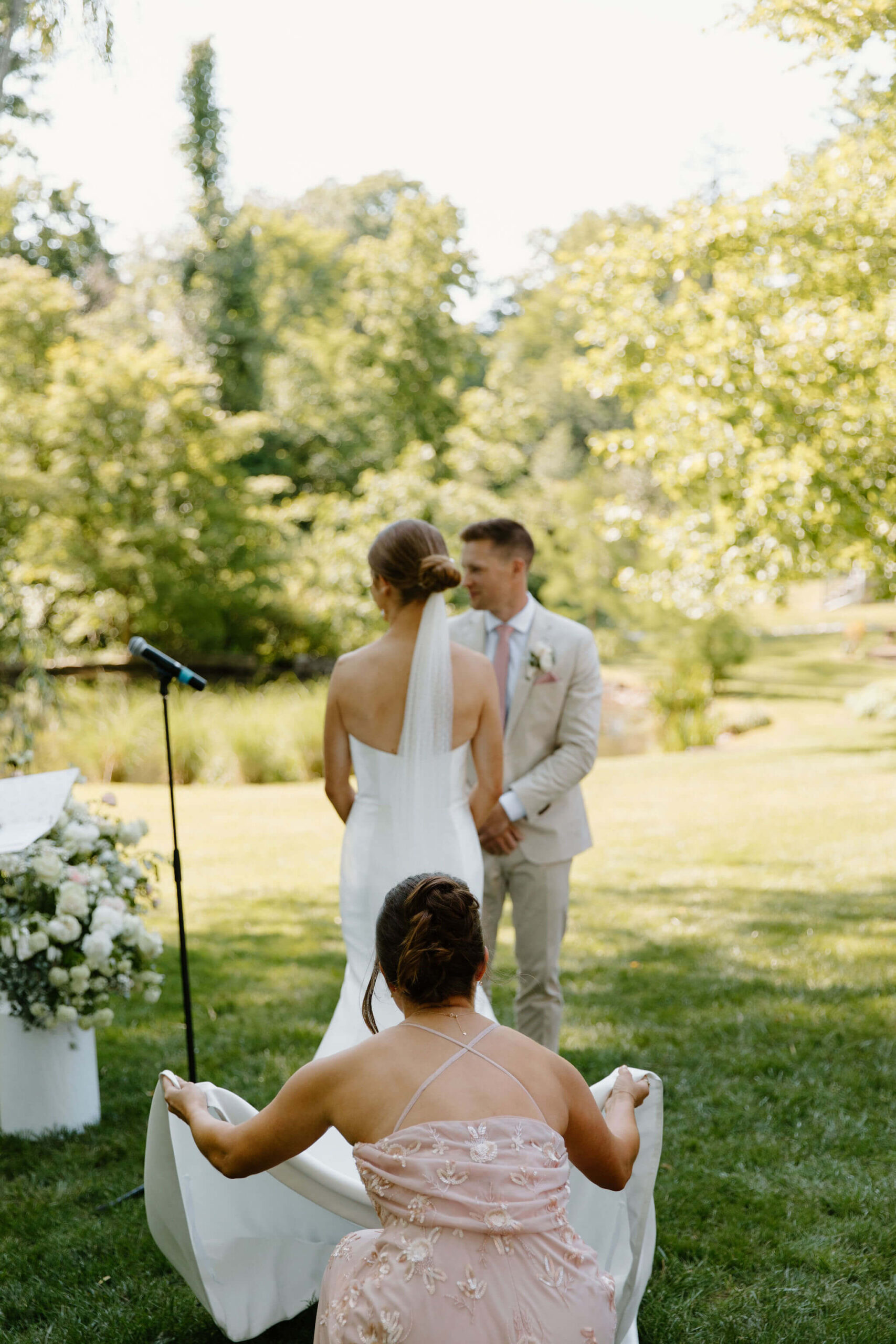 bride and groom standing at their wedding ceremony, bride's sister (her Maid of Honor) kneeling to straighten her train