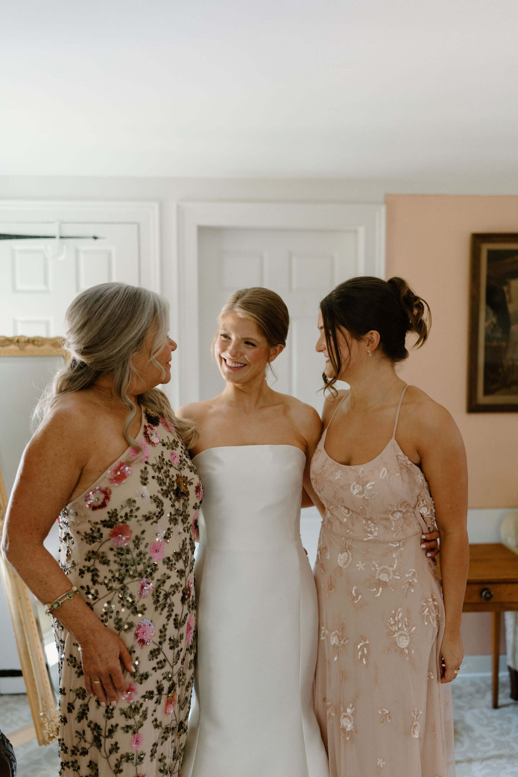 bride in a white wedding gown smiling at her mother (sequined floral embroidery) and sister (light pink gown with floral embroidery)