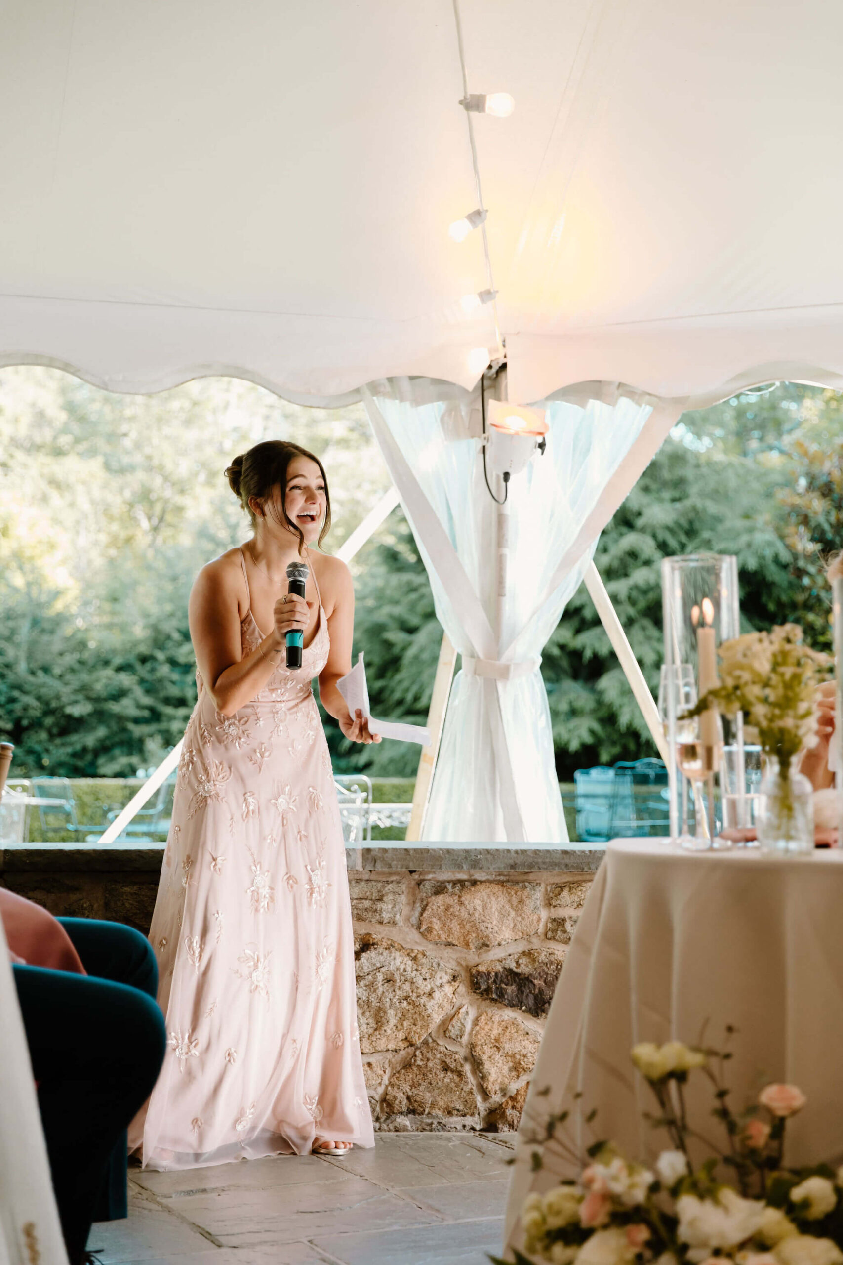 maid of honor (sister of the bride) wearing a pink dress with floral embroidery, laughing as she gives a wedding toast