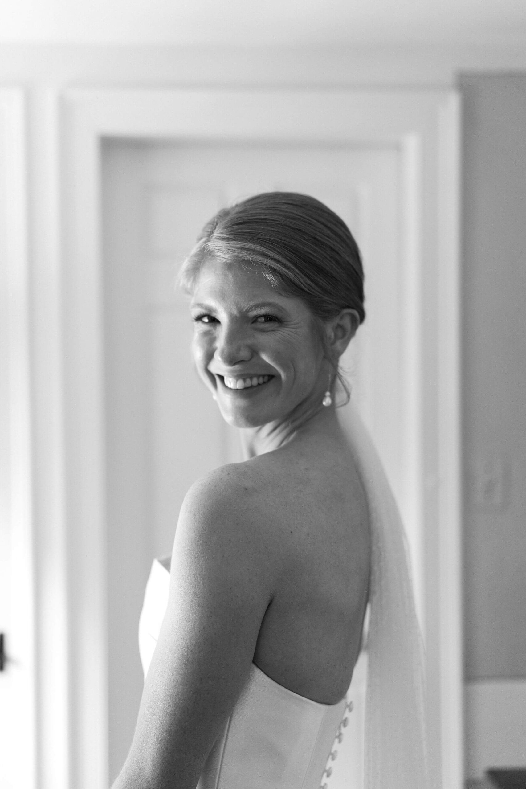 black and white image of a bride in her gown and veil, looking over her shoulder and smiling toward the camera