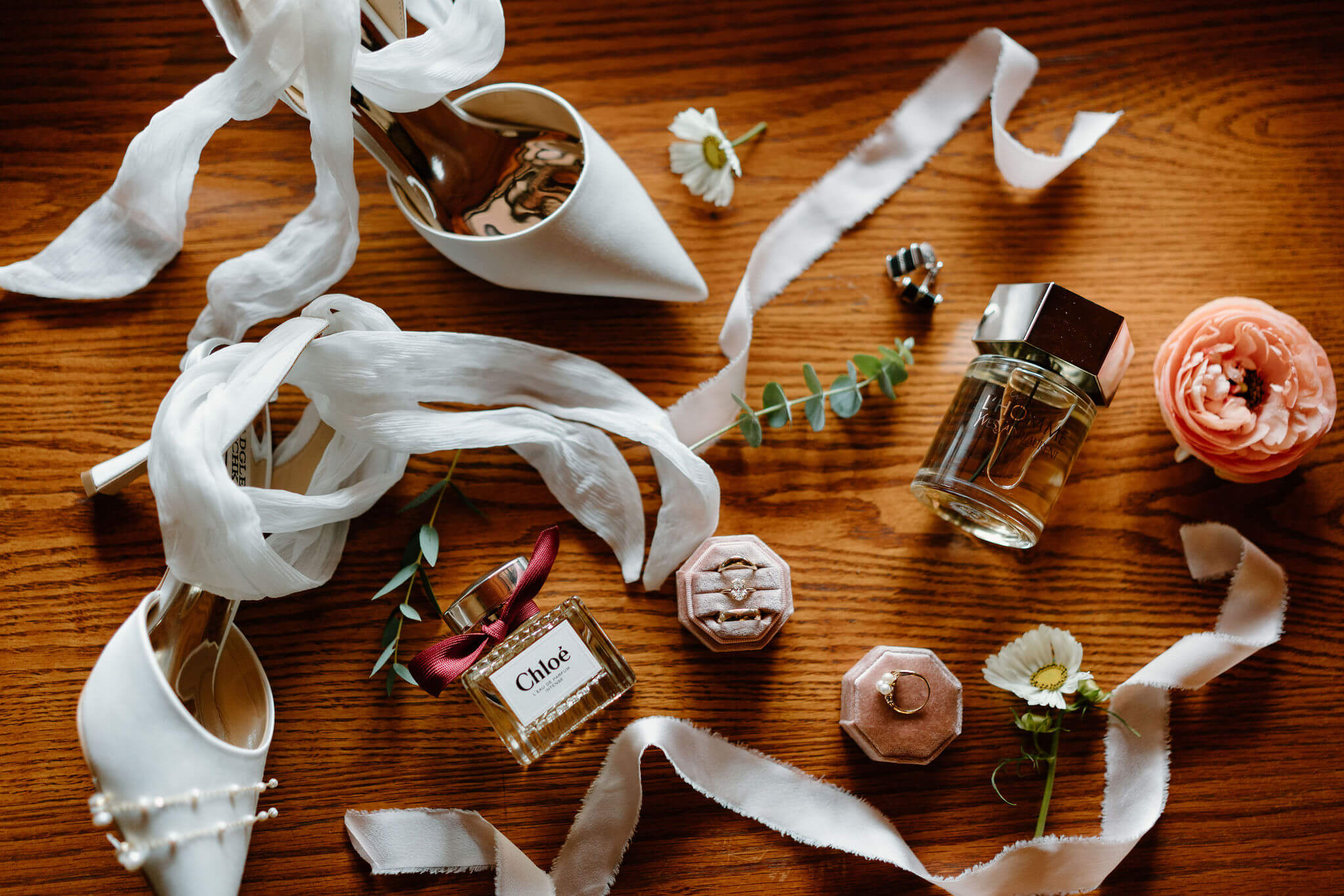 wedding flat lay featuring white bridal heels with fabric ties, perfume and cologne, two wedding bands, an engagement ring, and accents of ranunculus and eucalyptus