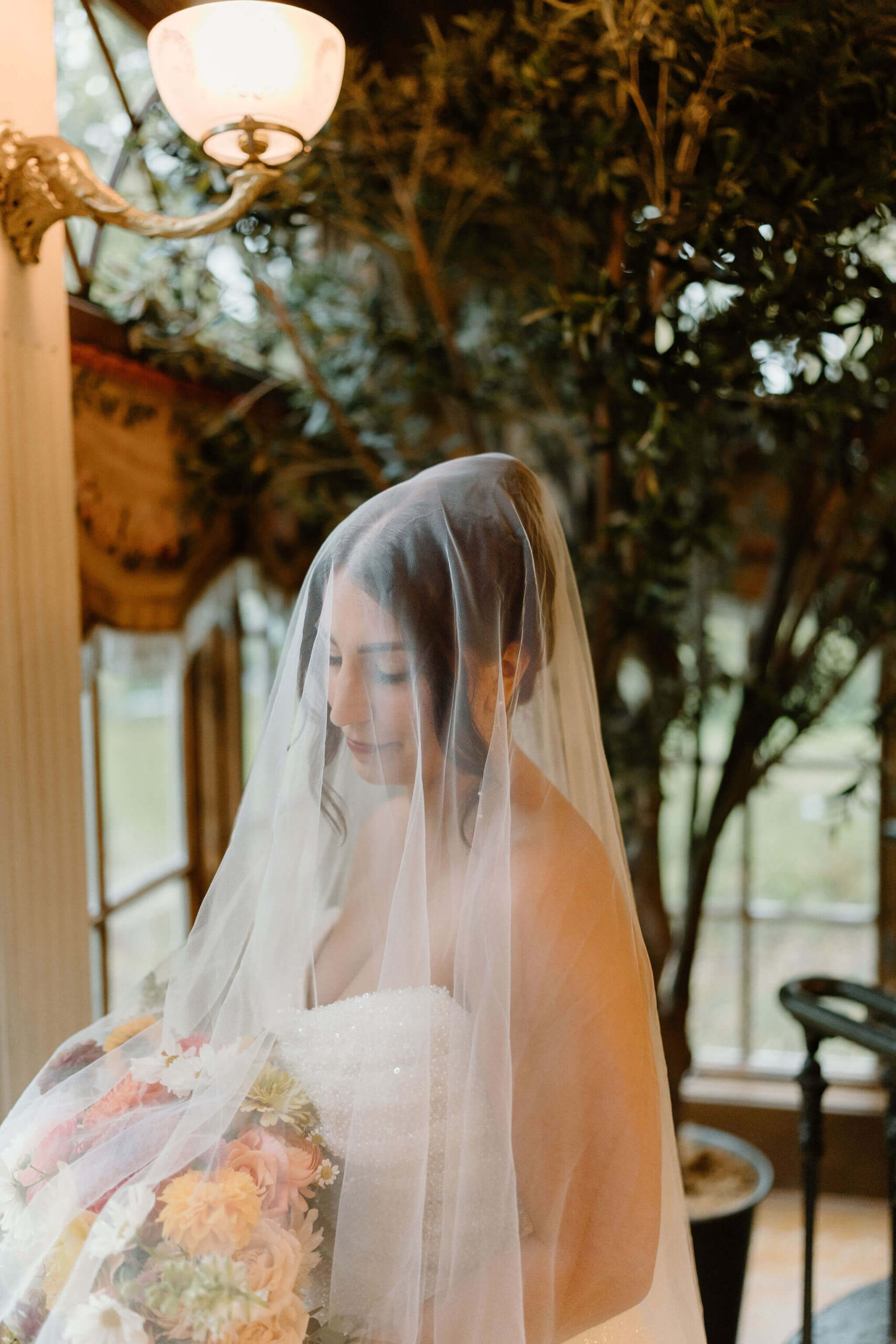 bride indoors against a window, smiling softly down at her flowers with her veil pulled over her face and draping down the flowers