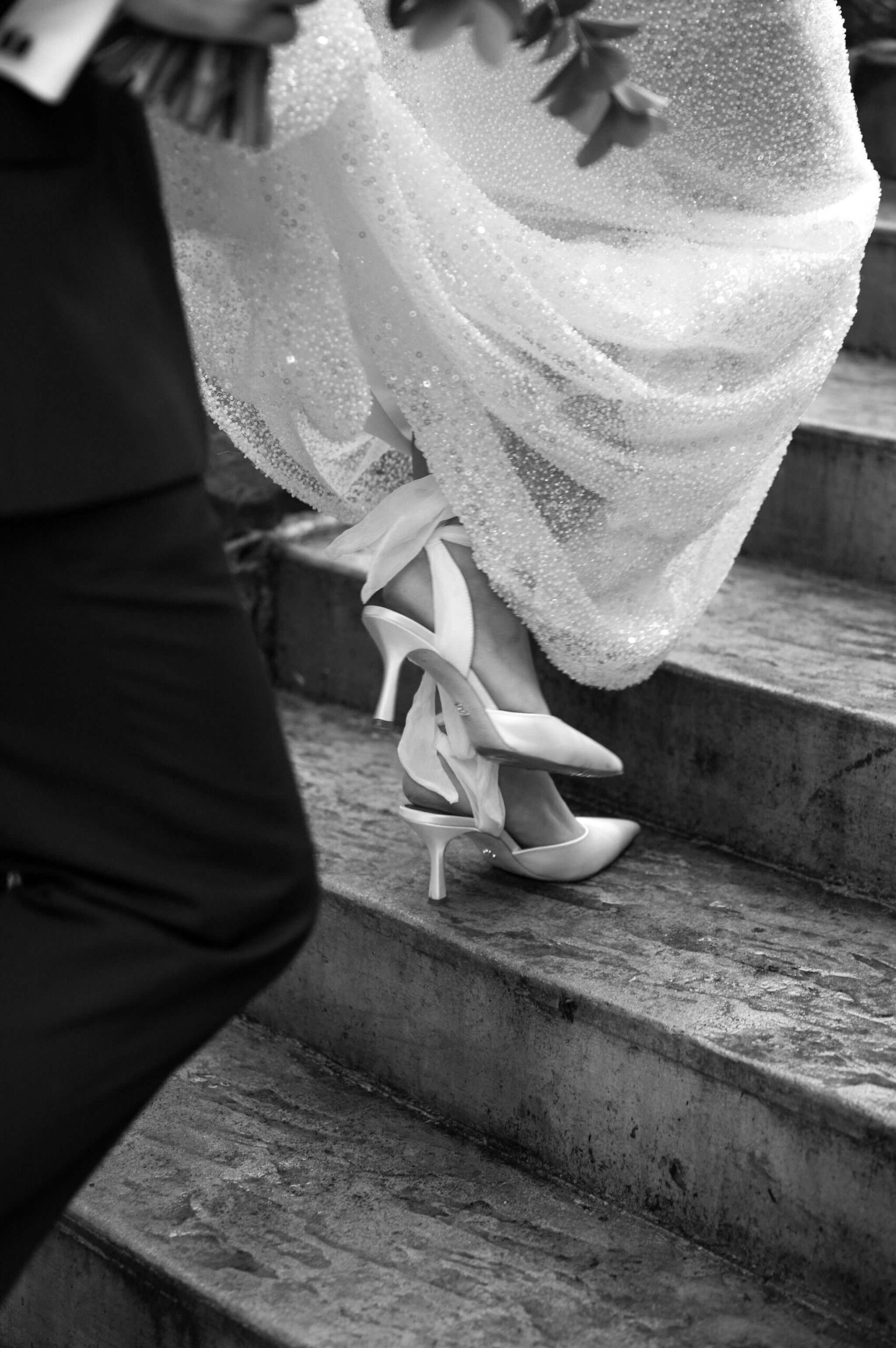 black and white close up of bride's shoes as she walks up a set of stairs, groom holding her bouquet in one hand and train of her dress in the other