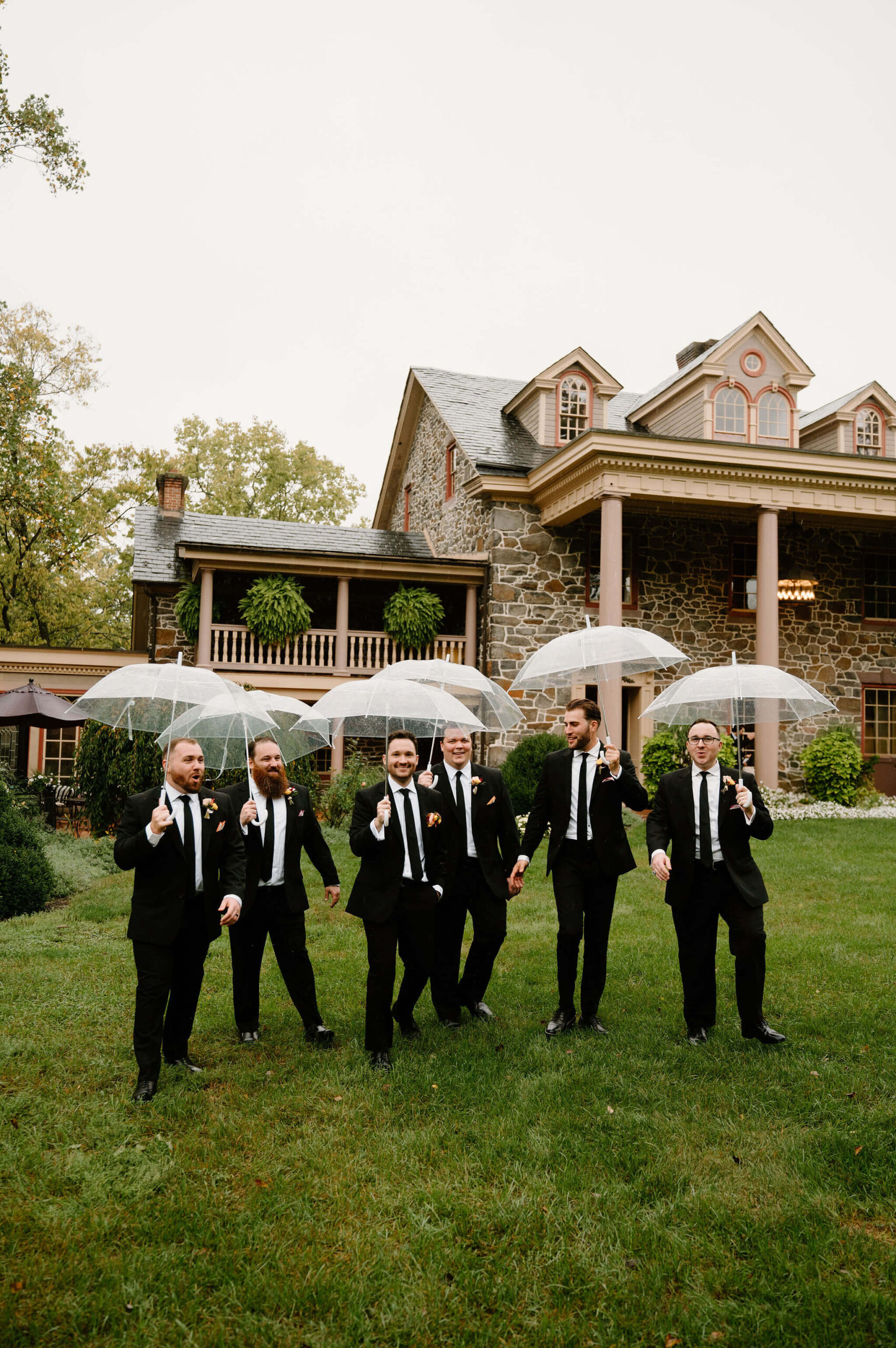 groom and groomsmen walking on the grass in front of a large stone estate, carrying clear umbrellas in the rain and laughing