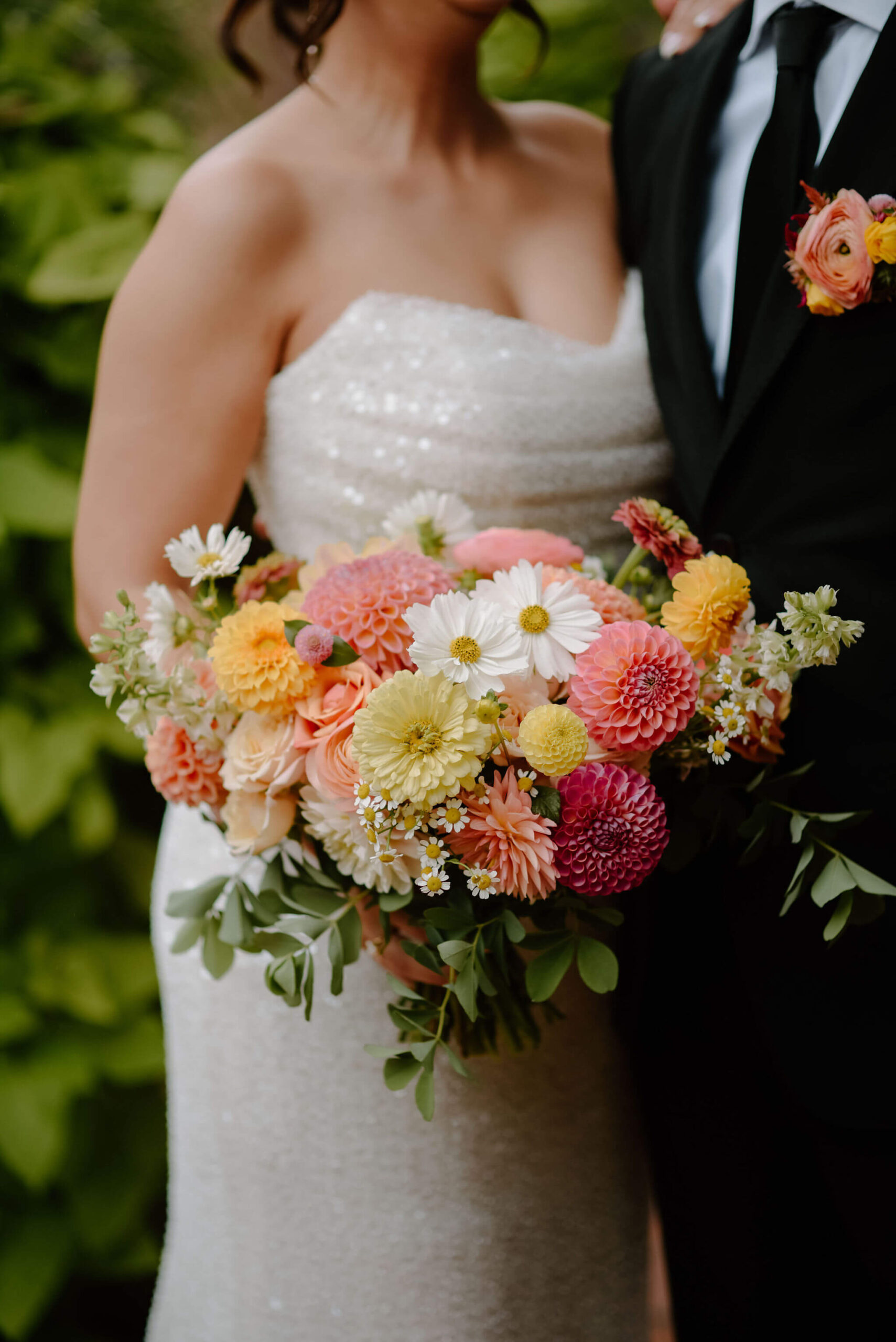 close up image of colorful pink, yellow, white, and greenery bridal bouquet, against a backdrop of the bride's sparkling wedding gown and groom's black suit with a colorful boutonniere