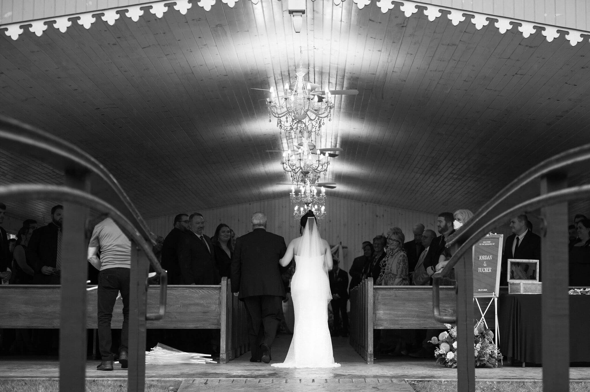 black and white image of bride and her father walking down the aisle at her wedding ceremony, from behind