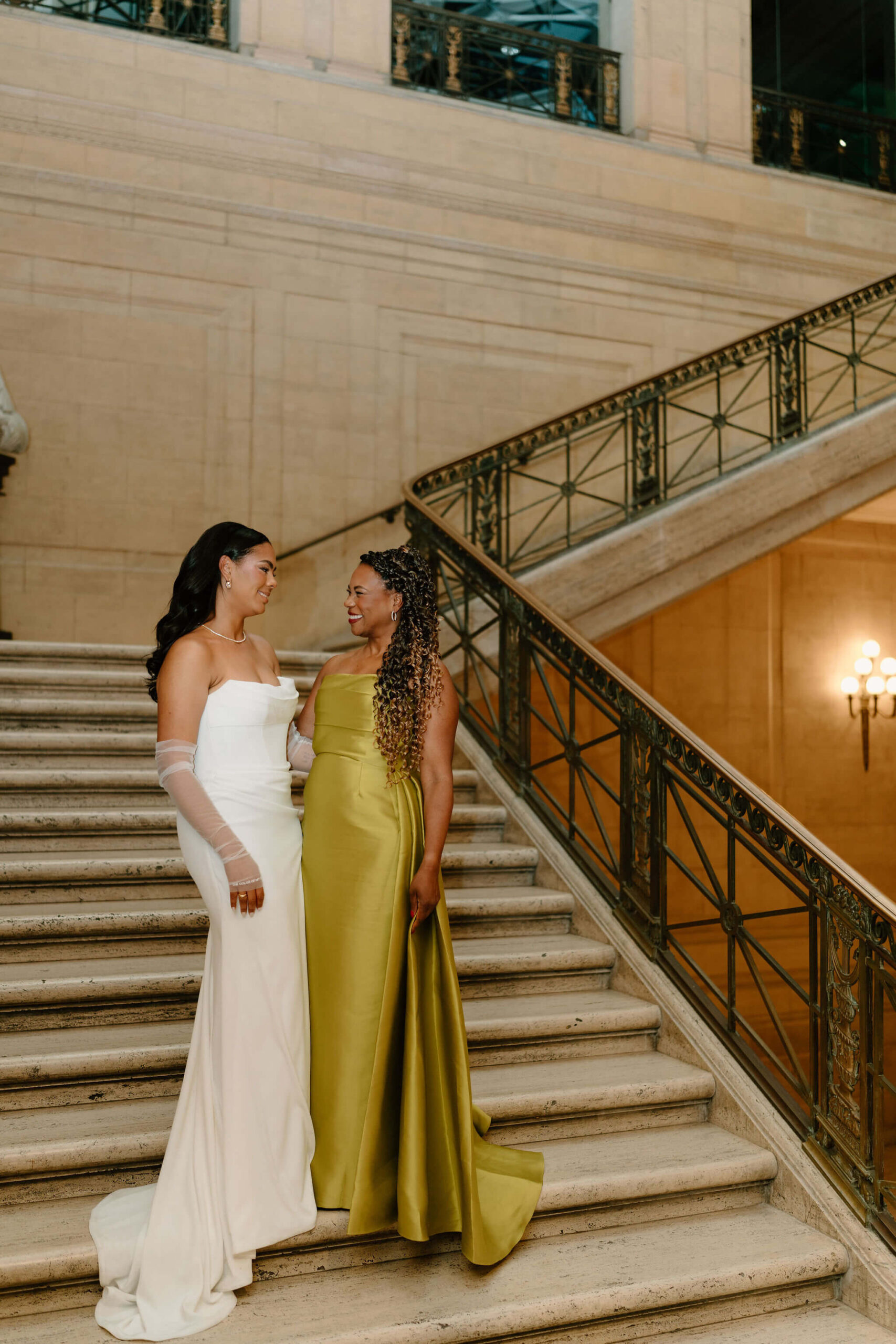 bride (white wedding gown) and her mother (chartreuse gown) smiling at each other on a marble staircase at her franklin institute wedding