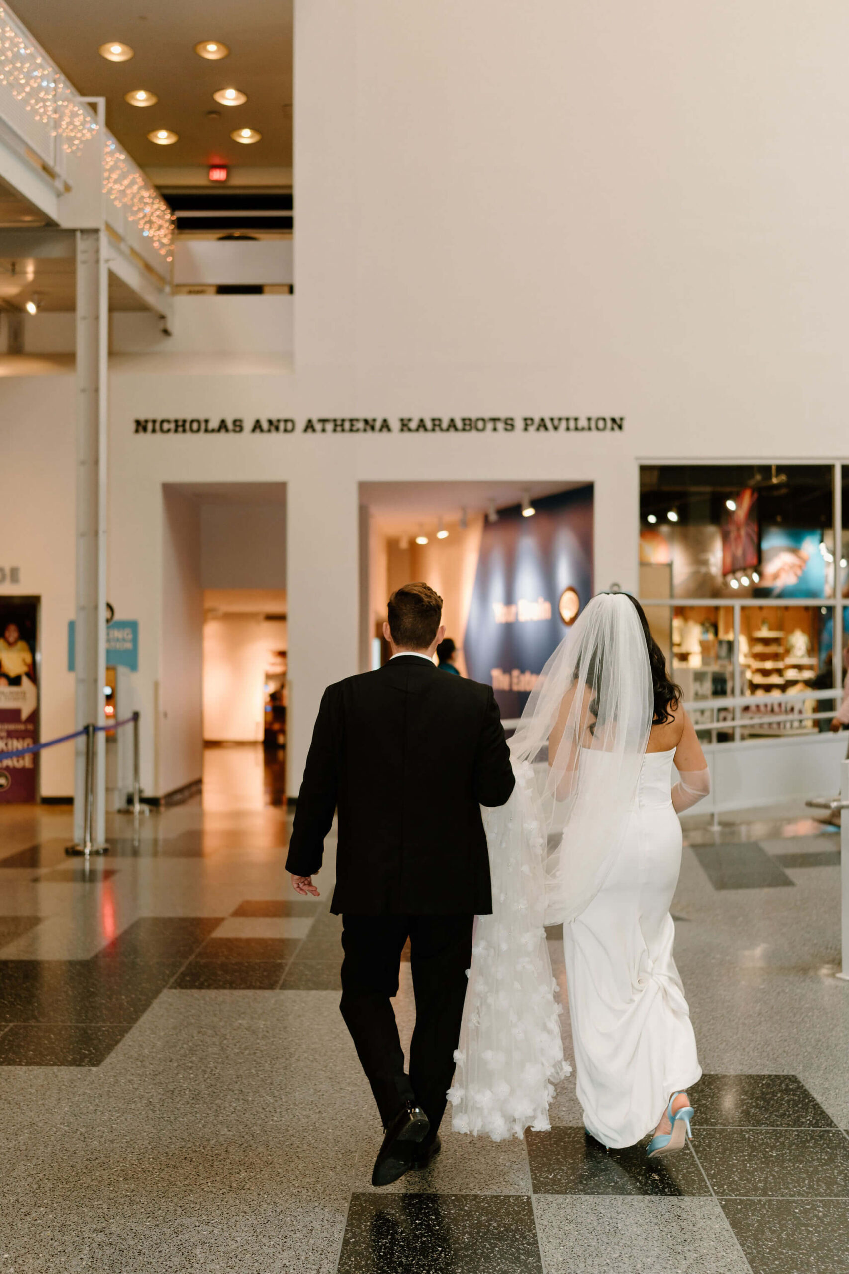 bride and groom walking through the lobby of the franklin institute together, towards a display labeled "Nicholas and Athena Karabots Pavilion"