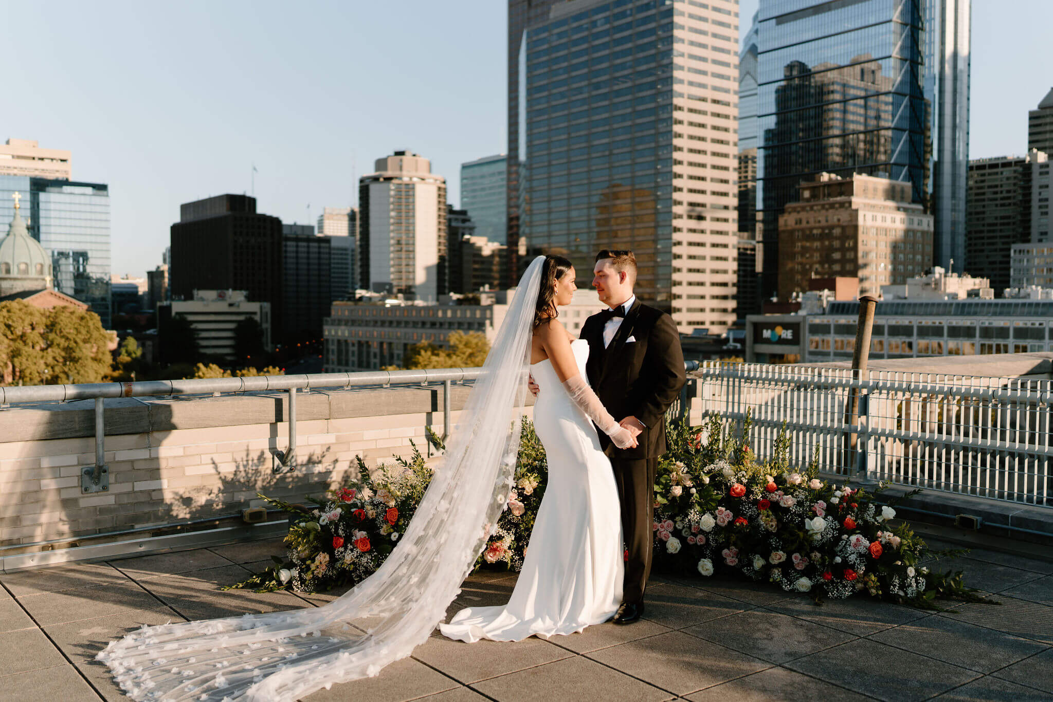 bride and groom looking softly at each other on the roof of the franklin institute, a floral installation on the ground behind them and skyscrapers rising in the distance, with the bride's veil flowing behind her dress at their franklin institute wedding