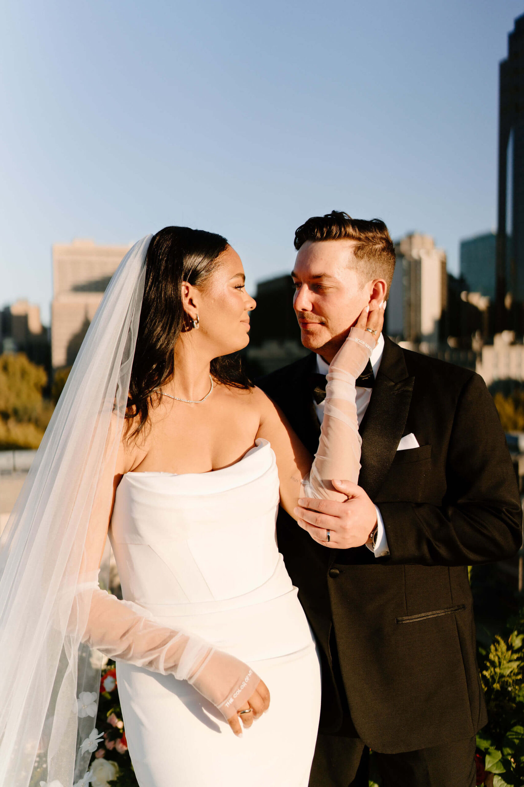 close-up of bride and groom looking at each other and smiling while standing side by side on the roof of the Franklin Institute, her left hand reaching up to hold his face and his left hand on her forearm