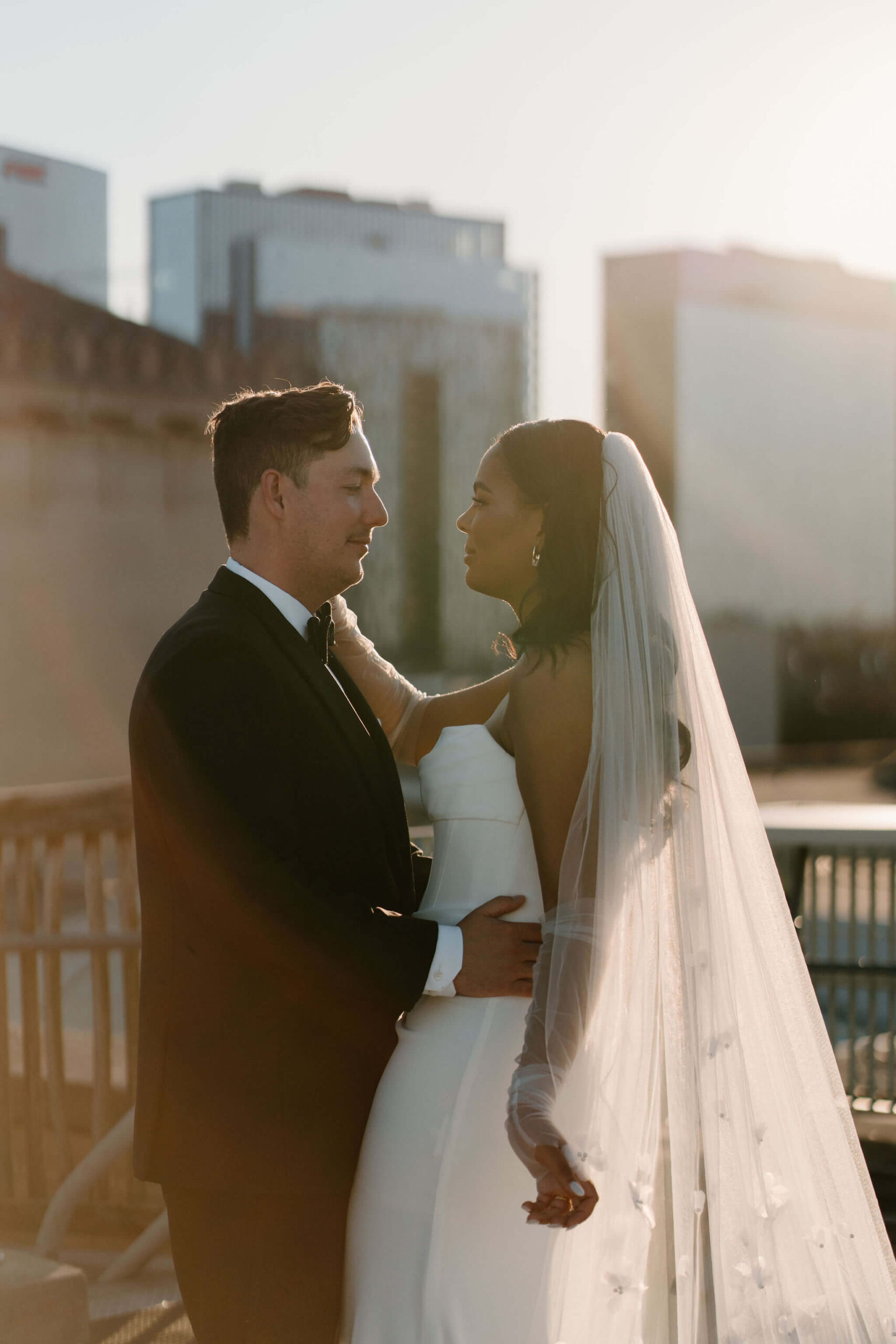bride and groom, backlit by the sun, looking softly at each other while the bride holds a corner of her veil