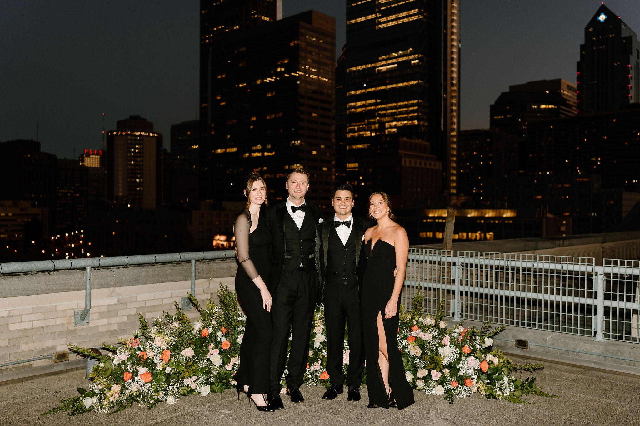 a group of friends, all wearing black, smiling at each with the lights of the Philadelphia skyline at dusk behind them