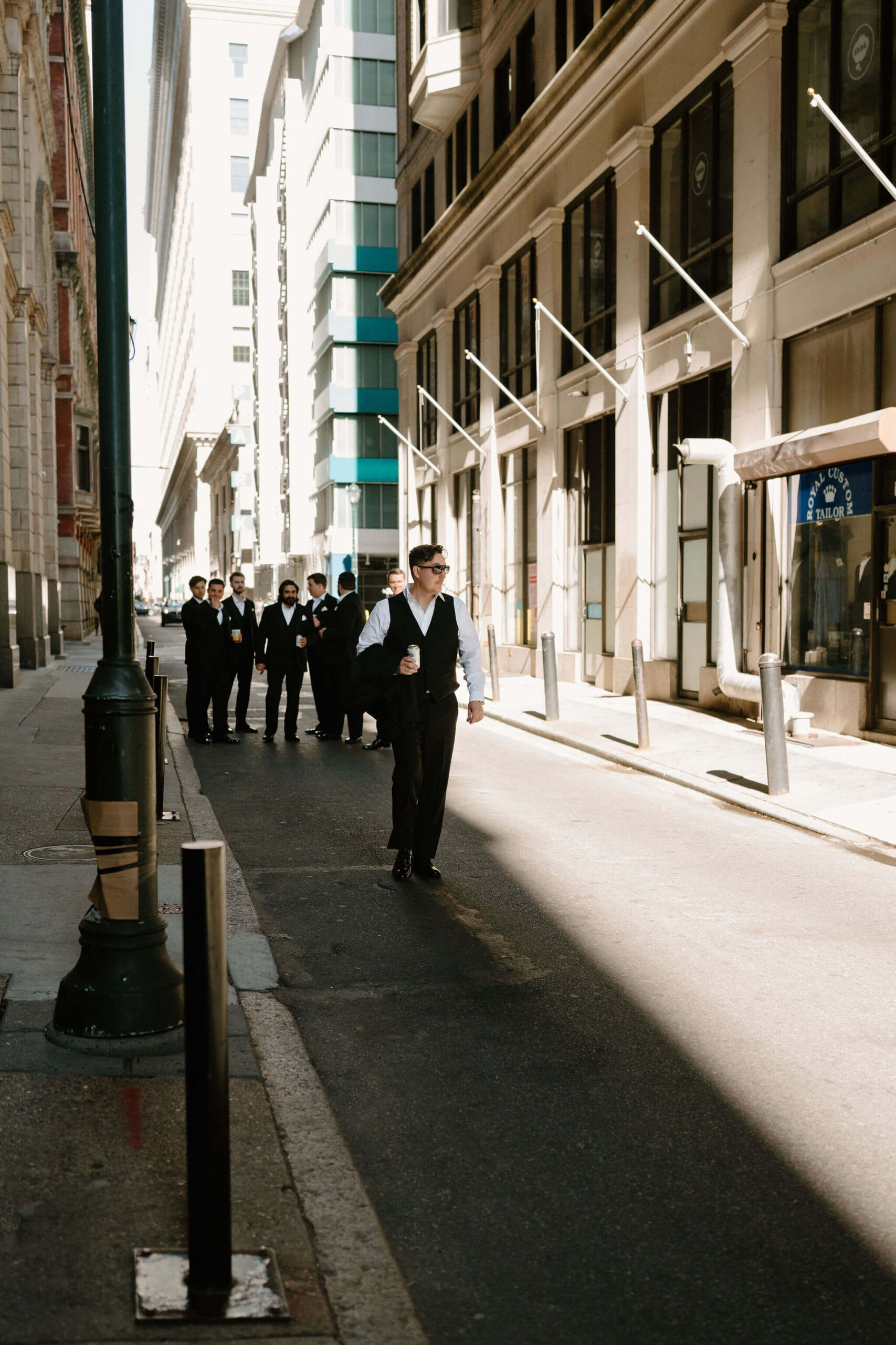 groom holding his black suitjacket in his hand and walking down a street in Philadelphia, looking off-camera, while his groomsmen stand in a group behind him (the kids call this "game day photos" like a sports team)