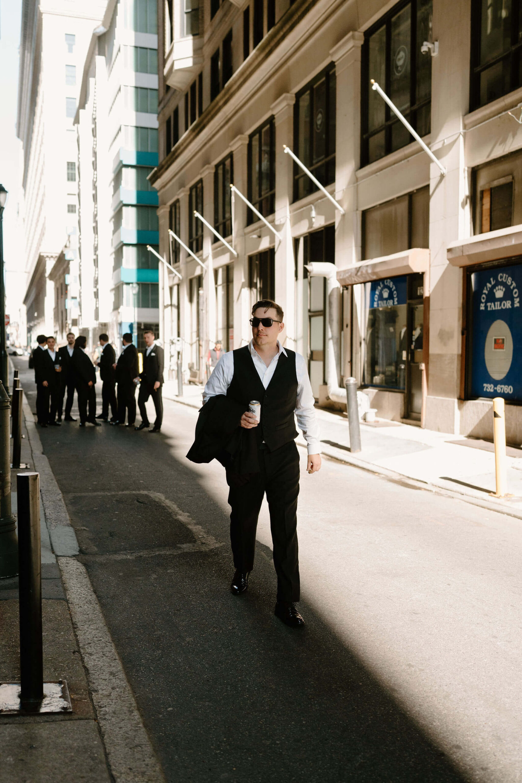 groom holding his black suit jacket in his hand, as well as a beer, and walking down a street in Philadelphia, looking off-camera, while his groomsmen stand in a group behind him