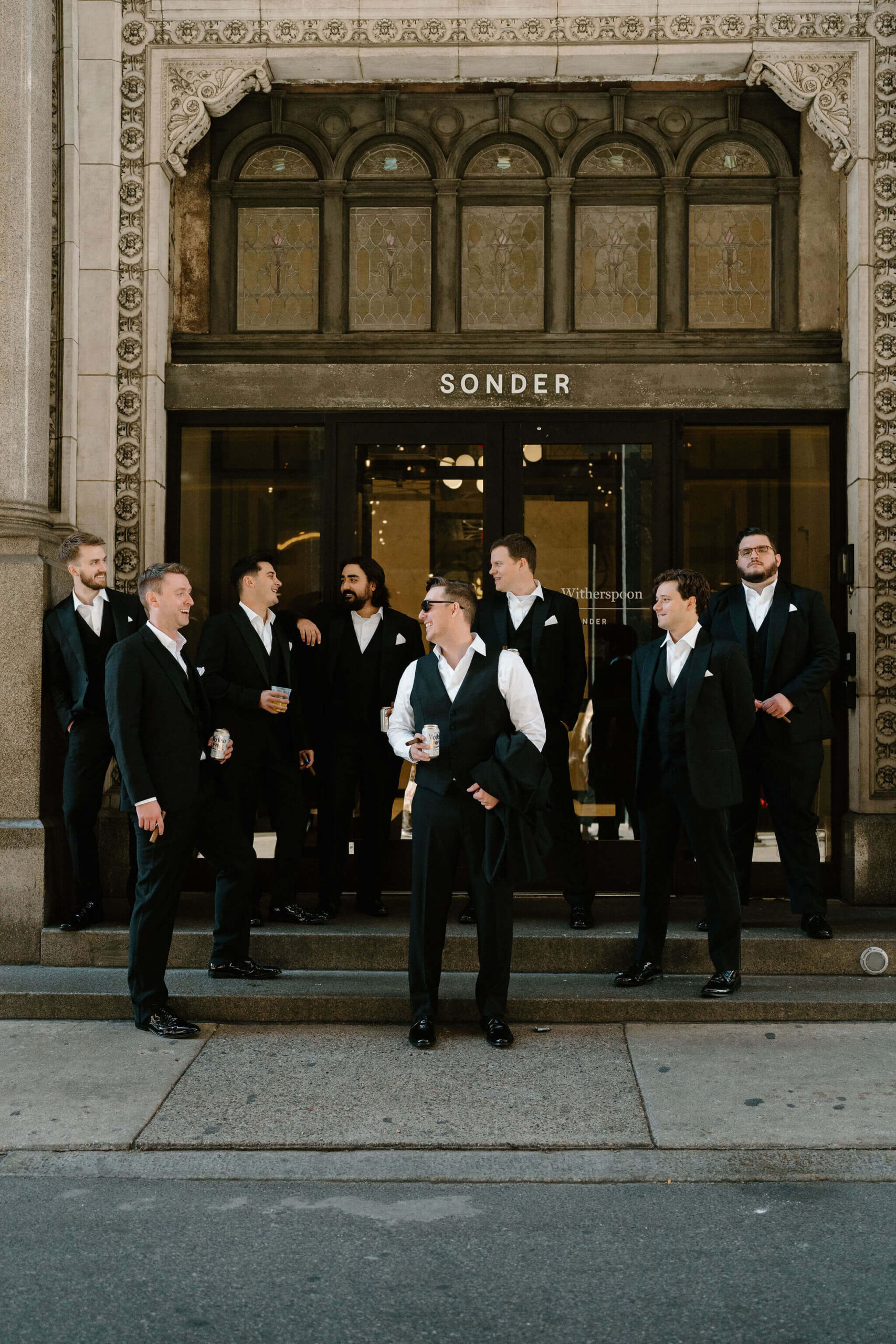 groom and groomsmen in black suits standing on the steps in front of a building with a sign that reads "Sonder", looking at each other and talking