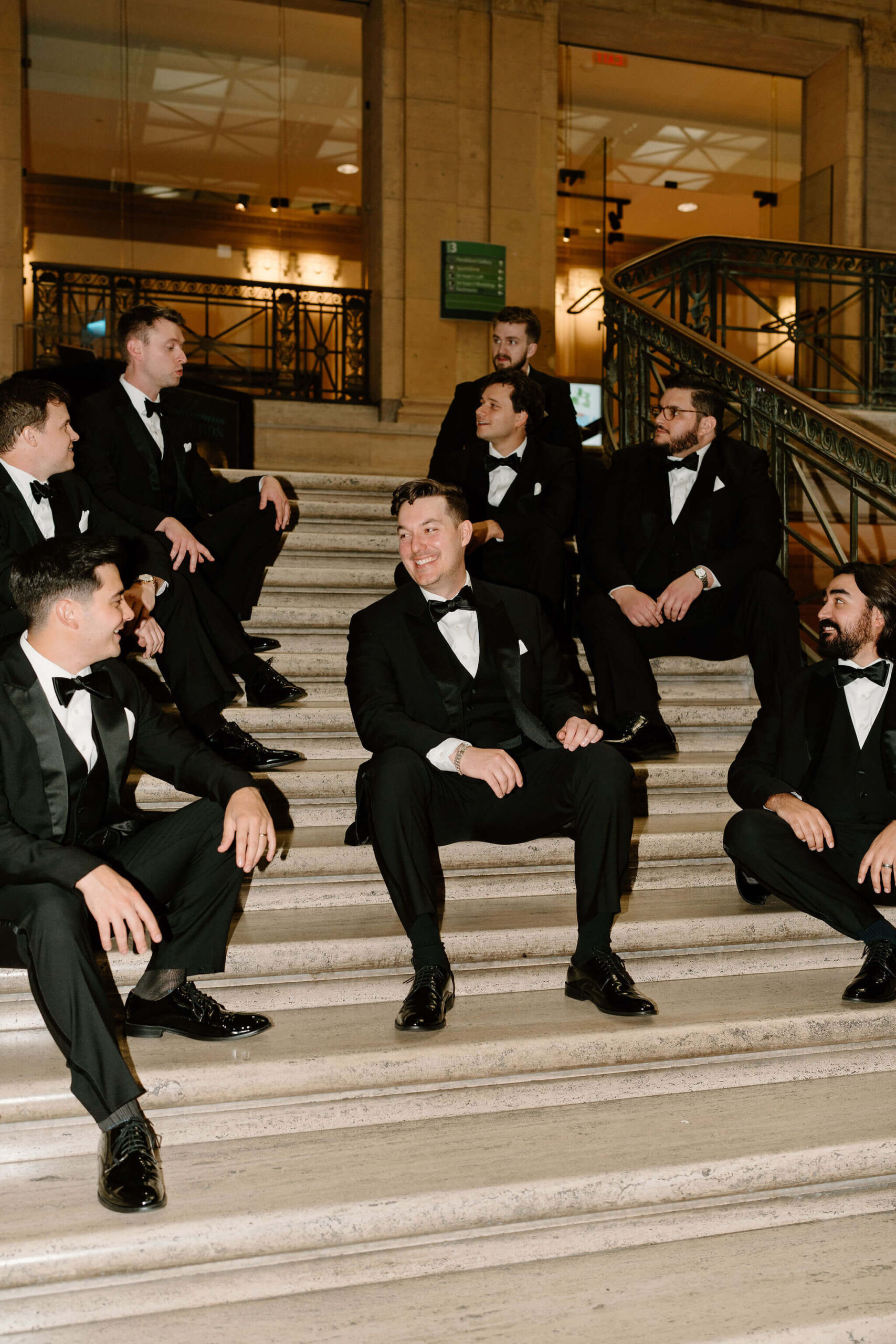 groom and groomsmen, all wearing black tuxedos, seated on a marble staircase and all smiling at each other at his franklin institute wedding