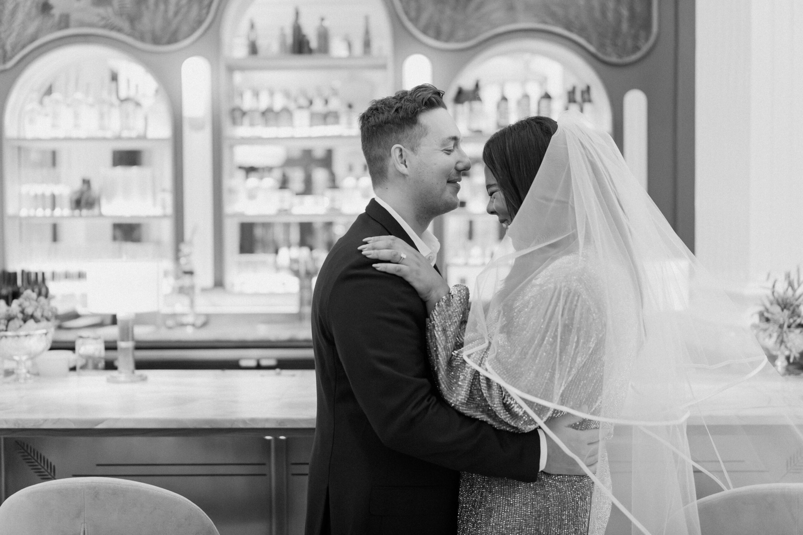 black and white image of an engaged couple embracing in front of a beautiful cocktail bar, the bride wearing a tulle veil, and smiling at each other