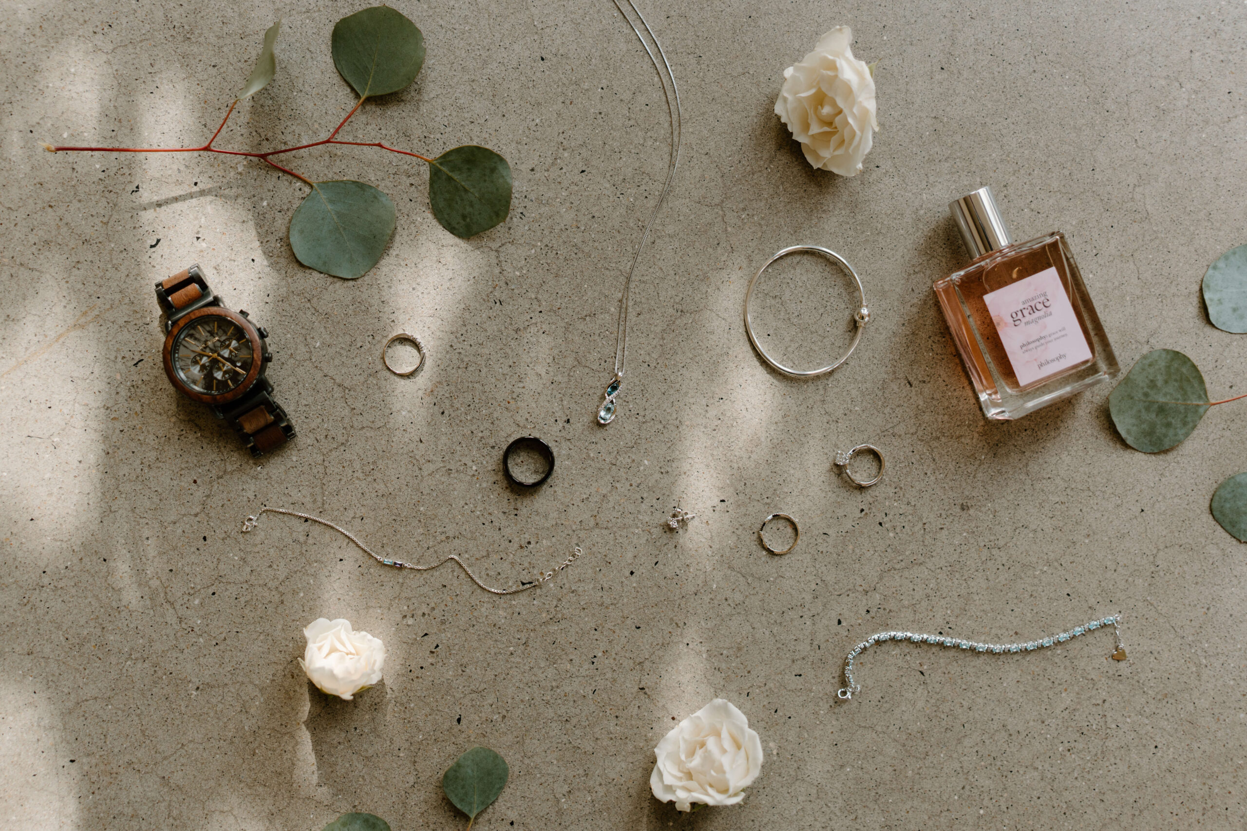 wedding flat-lay featuring groom's watch and wedding band, with bride's necklace, bracelets, earrings, engagement ring, wedding band, and perfume, surrounded by florals and eucalyptus