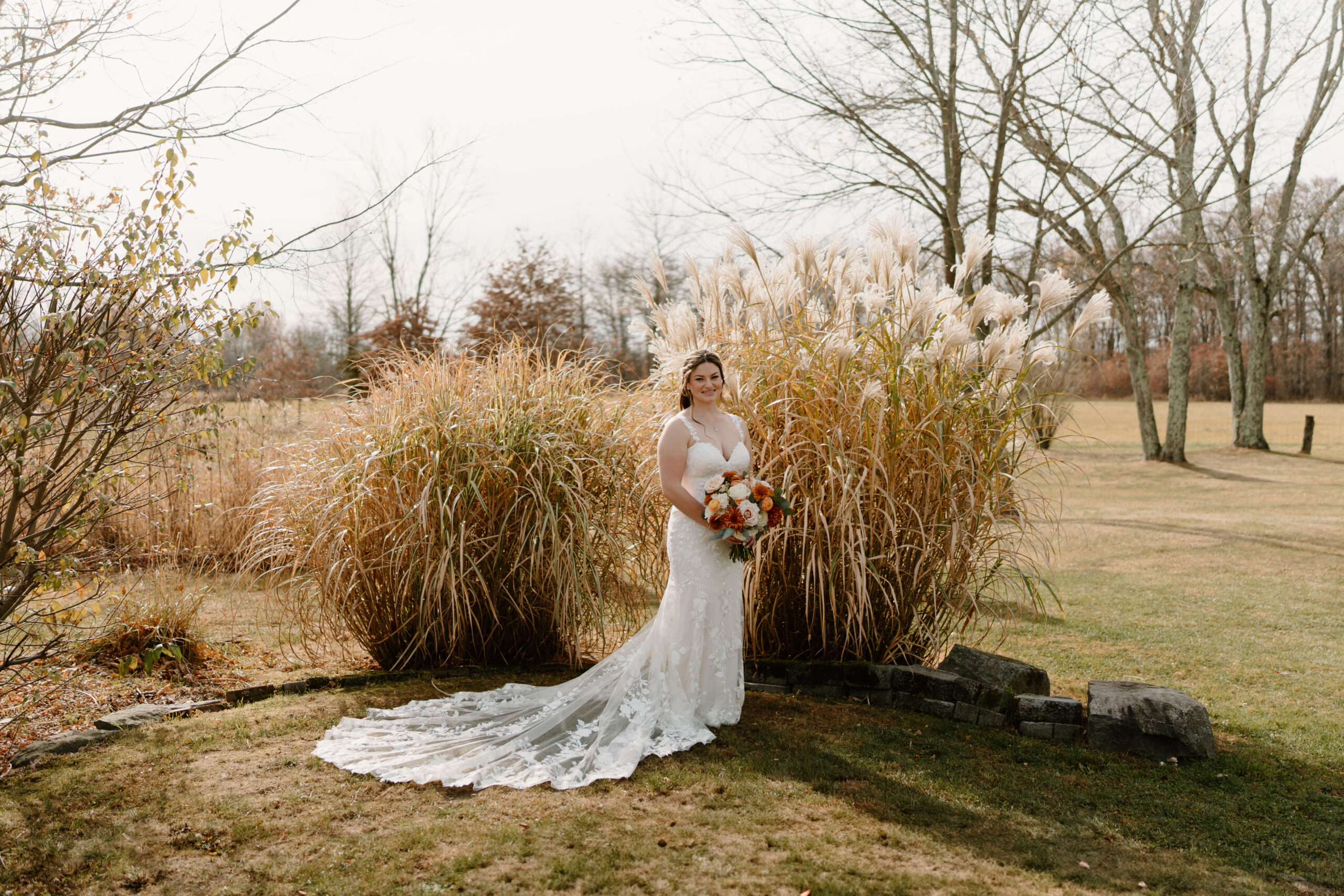 bride in beautiful lace wedding dress with train extending behind her, holding a large bouquet of orange, white, and red flowers, smiling at the camera