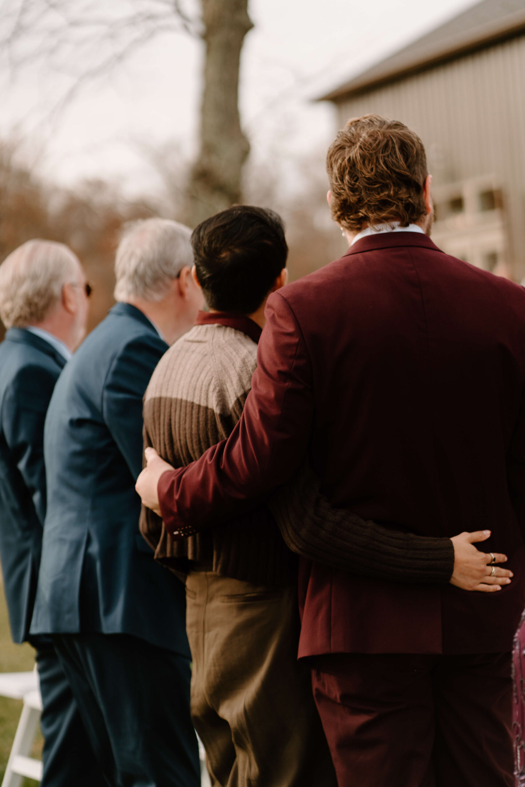 brother of the bride and his boyfriend embracing, while watching bride walk down the aisle at her wedding ceremony