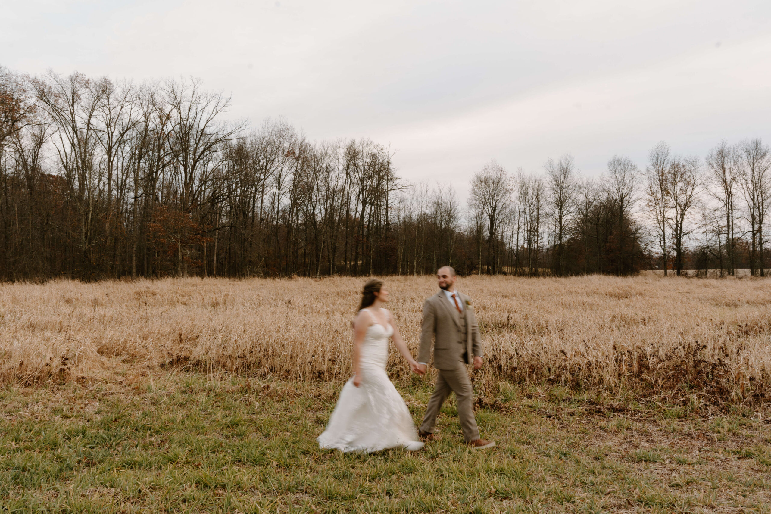 motion-blur image of bride and groom holding hands and walking through a field at sunset