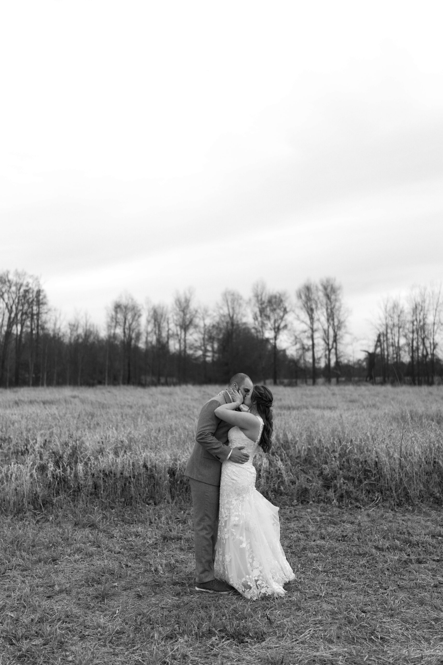 black and white image of bride holding groom's face and kissing him in a field on wedding day