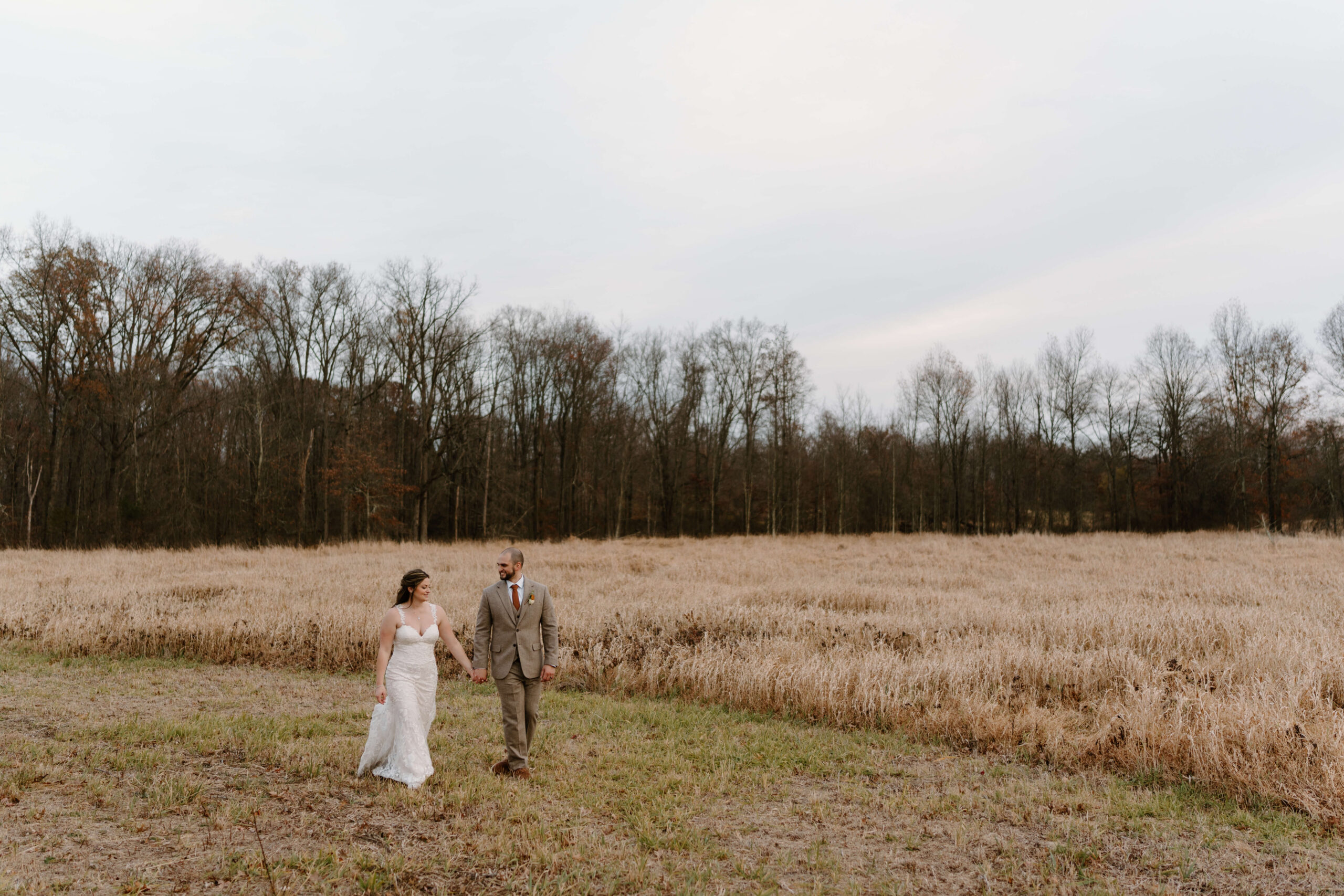bride and groom holding hands and walking through a field at sunset in November on their wedding day at the Farm Bakery and Events
