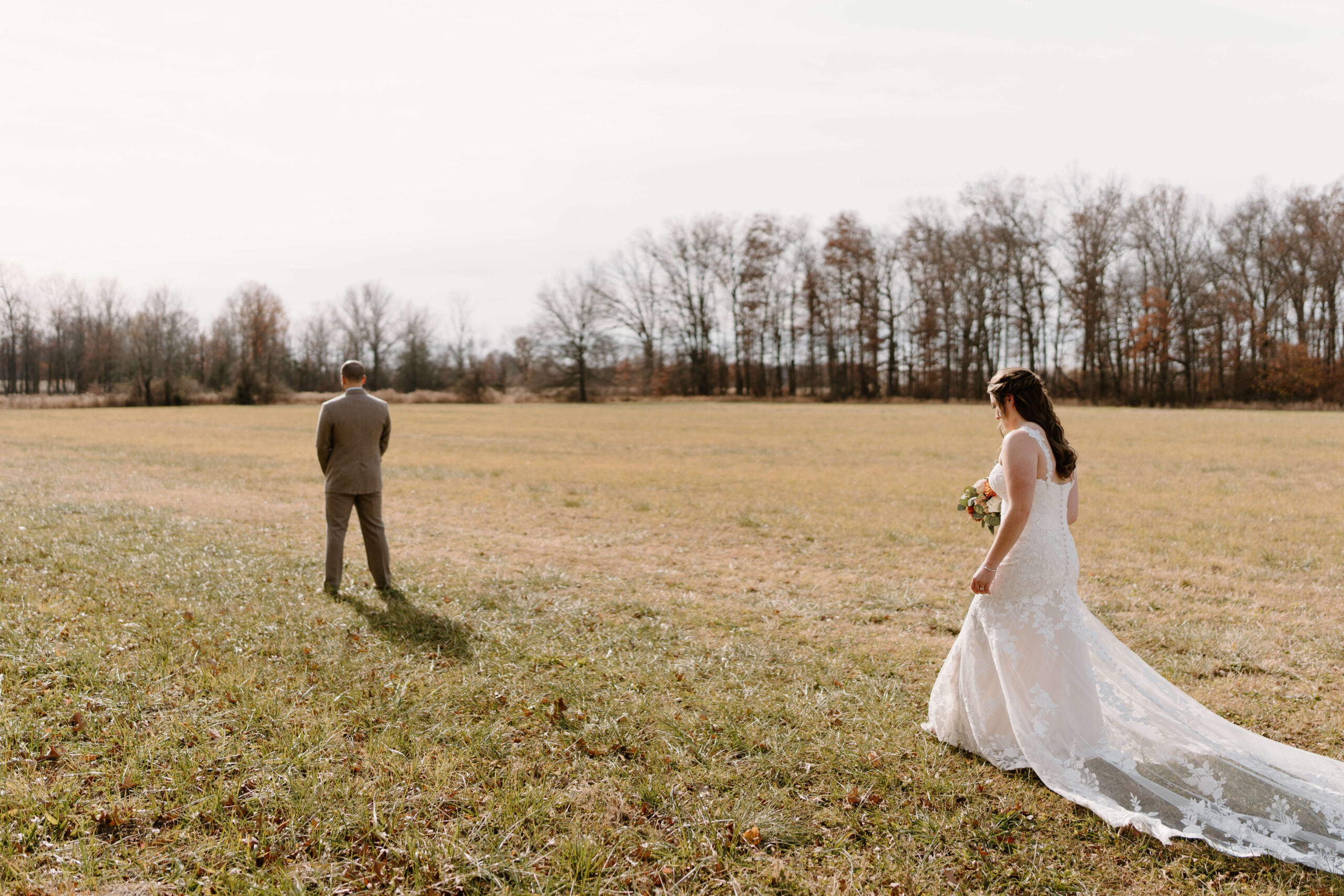 bride approaching groom in an open field, groom facing away from her, in preparation for their first look on their wedding day